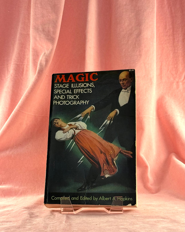 Book titled 'Magic: Stage Illusions, Special Effects and Trick Photography' on a stand with a pink curtain background