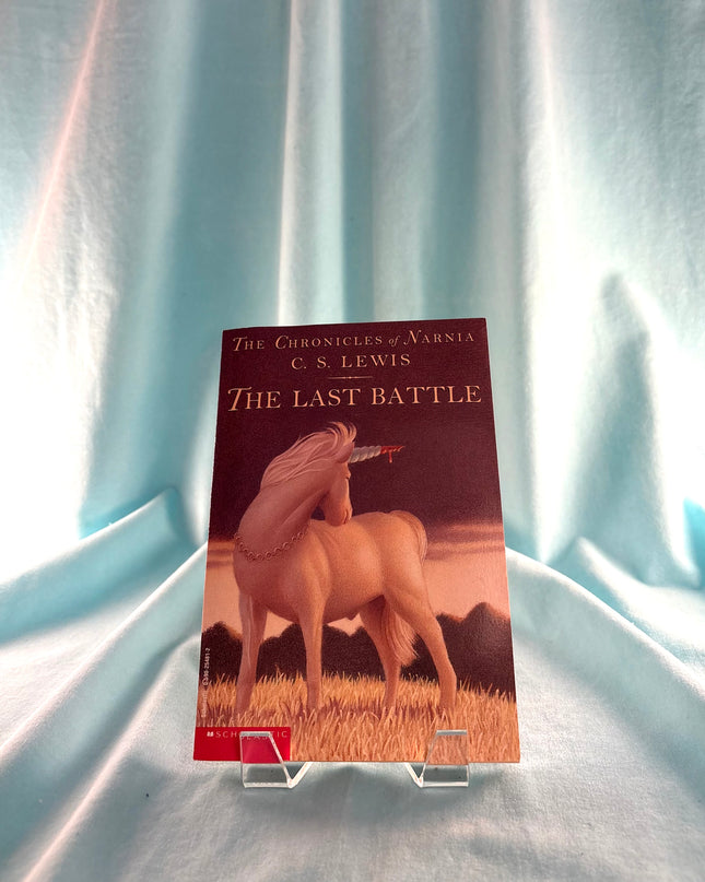 Book cover of 'The Last Battle' from 'The Chronicles of Narnia' series on a stand against a light blue curtain background.