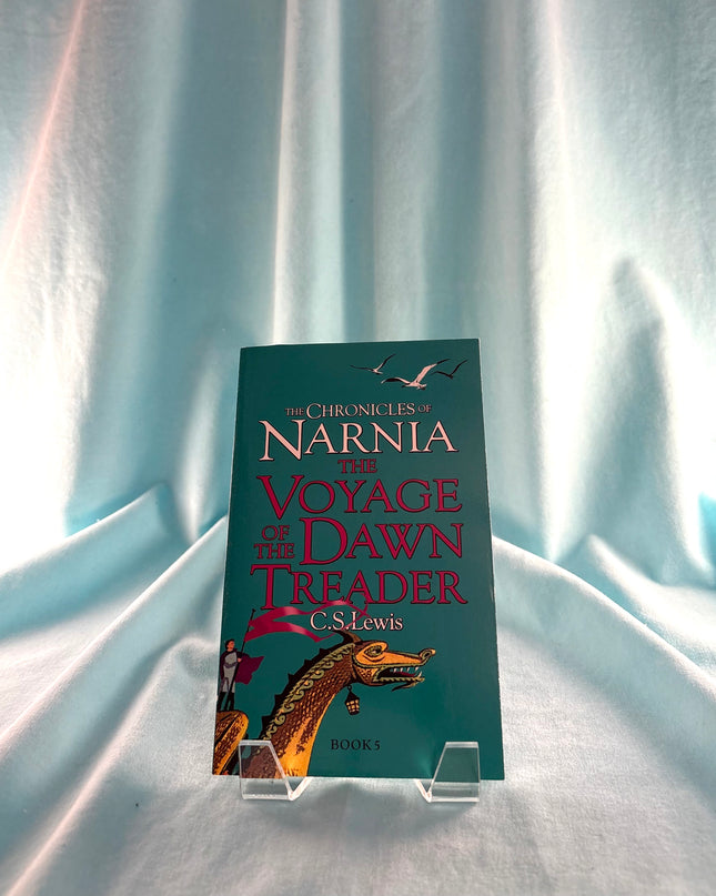 Book titled 'The Voyage of the Dawn Treader' from 'The Chronicles of Narnia' series on a stand against a light curtain background.