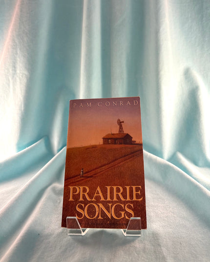 Book titled 'Prairie Songs' by Pam Conrad on a stand against a light blue curtain background