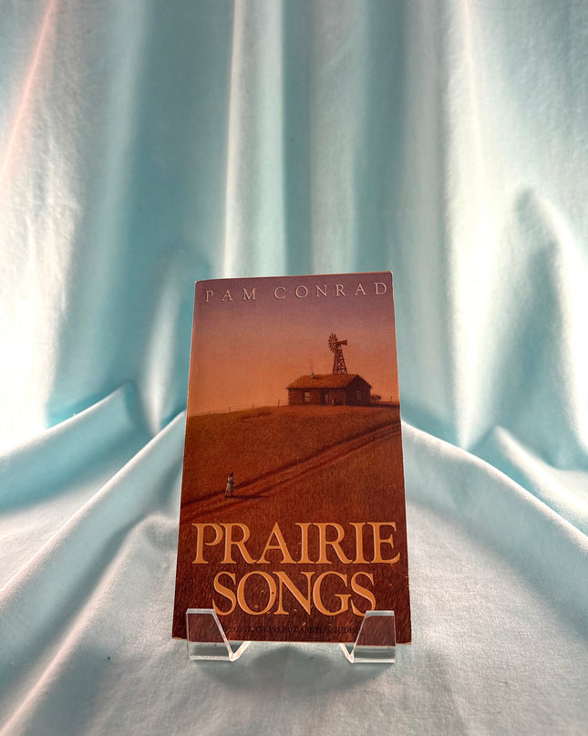 Book titled 'Prairie Songs' by Pam Conrad on a stand against a light blue curtain background