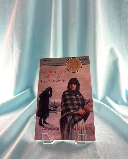 Book titled 'A Gathering of Days' on a stand with a light blue curtain background