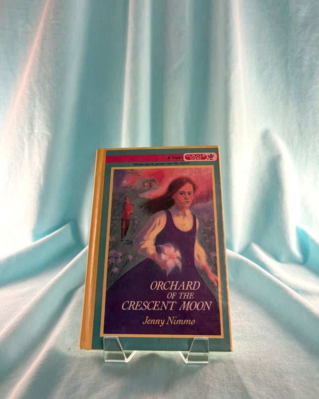 Book titled 'Orchard of the Crescent Moon' by Jenny Nimmo on a stand against a light curtain background