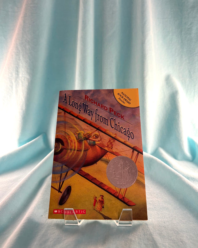 Book titled 'A Long Way from Chicago' by Richard Peck on a clear stand against a light blue curtain background.