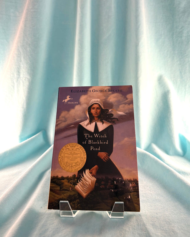 Book titled 'The Witch of Blackbird Pond' by Elizabeth George Speare on a stand against a light blue curtain background.