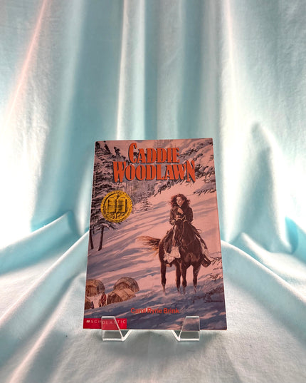 Book titled 'Caddie Woodlawn' on a stand against a light curtain background