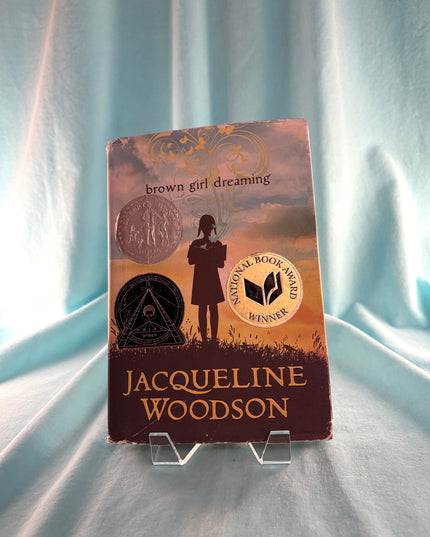Book titled 'Brown Girl Dreaming' by Jacqueline Woodson on a stand against a light blue curtain background