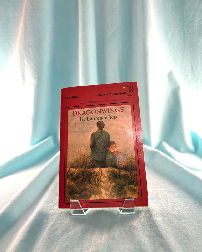 Book titled 'Dragonwings' by Laurence Yep on a clear stand against a light blue curtain background