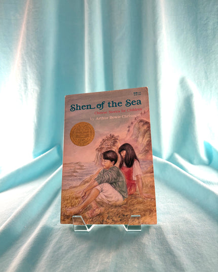 Book titled 'Shen of the Sea' on a stand with a light blue curtain background