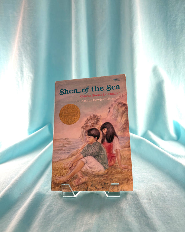 Book titled 'Shen of the Sea' on a stand with a light blue curtain background