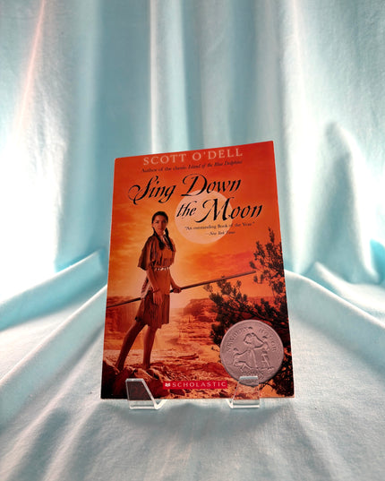 Book titled 'Sing Down the Moon' by Scott O'Dell on a clear stand against a light blue curtain background.