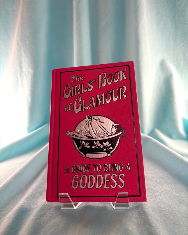 Red book titled 'The Girl's Book of Glamour' on a clear stand against a light blue curtain background