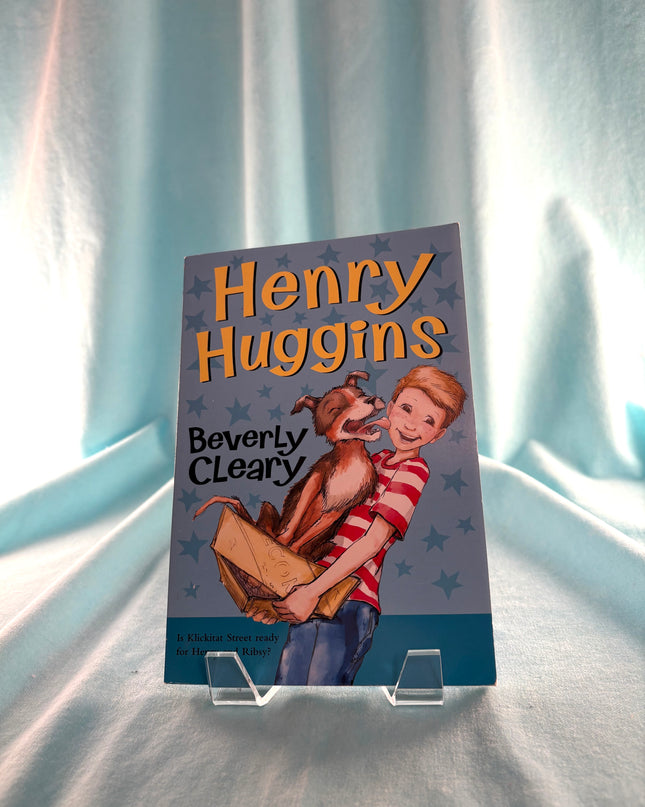 Book titled 'Henry Huggins' by Beverly Cleary on a stand with a light blue curtain background