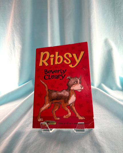 Book titled 'Ribsy' by Beverly Cleary on a clear stand against a light curtain background