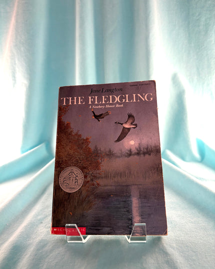 Book titled 'The Fledgling' on a stand with a light blue curtain background