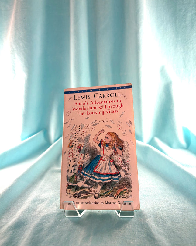 Book titled 'Alice's Adventures in Wonderland and Through the Looking Glass' by Lewis Carroll on a stand against a light blue curtain background.