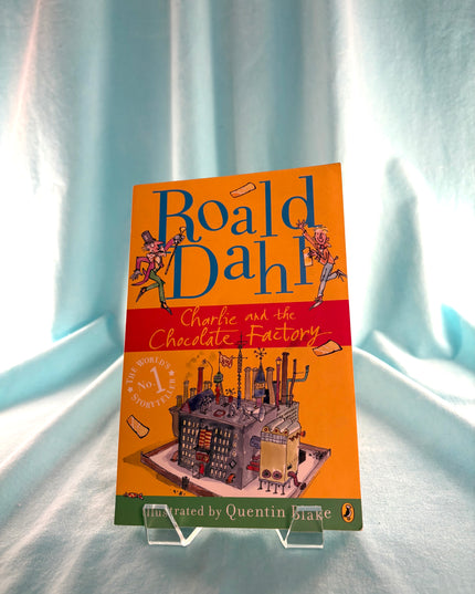 Book cover of 'Charlie and the Chocolate Factory' by Roald Dahl on a stand against a light curtain background