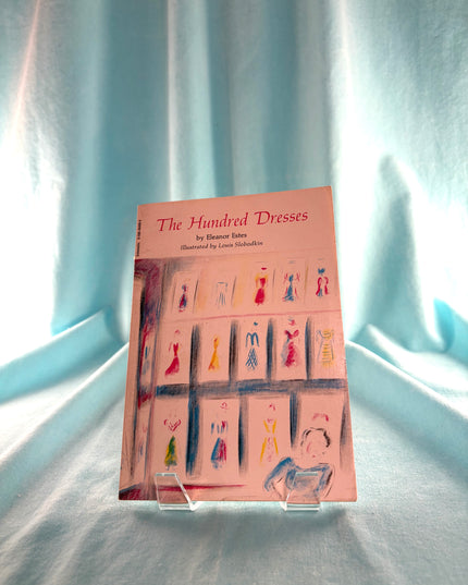 Book titled 'The Hundred Dresses' on a stand with a light blue curtain background