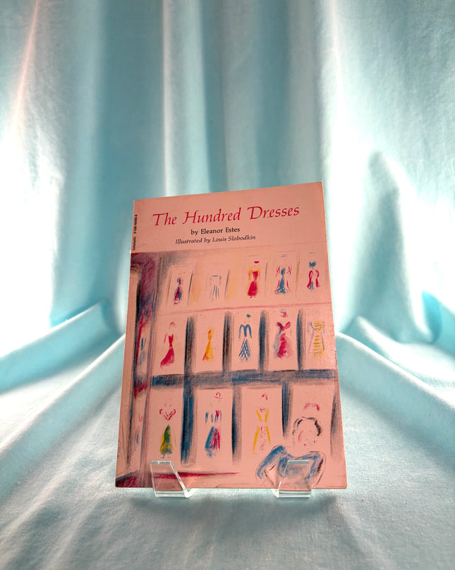Book titled 'The Hundred Dresses' on a stand with a light blue curtain background
