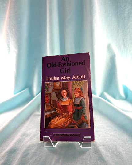Book titled 'An Old-Fashioned Girl' by Louisa May Alcott on a clear stand against a light curtain background