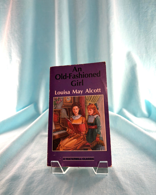 Book titled 'An Old-Fashioned Girl' by Louisa May Alcott on a clear stand against a light curtain background