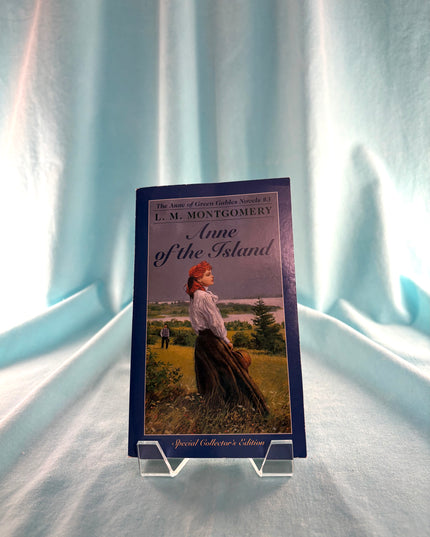 Book titled 'Anne of the Island' by L.M. Montgomery on a stand against a light blue curtain background