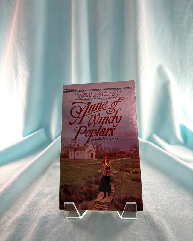 Book titled 'Anne of Windy Poplars' on a clear stand against a light blue curtain background