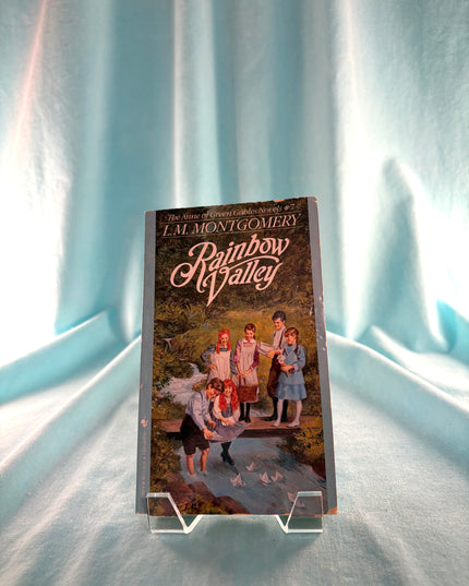 Book titled 'Rainbow Valley' by L.M. Montgomery on a stand against a light blue curtain background