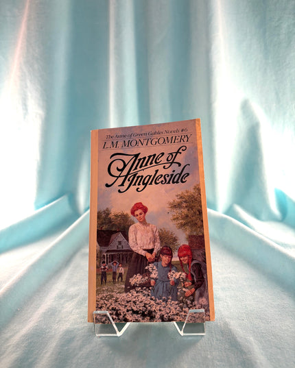 Book titled 'Anne of Ingleside' by L.M. Montgomery on a stand with a light blue curtain background