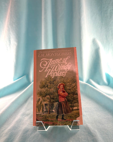 Book titled 'Anne of a Windy Poplar' by L.M. Montgomery on a stand against a light blue curtain background.