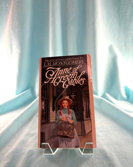 Book titled 'Anne of Green Gables' by L.M. Montgomery on a stand against a light blue curtain background.