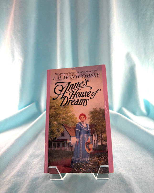 Book titled 'Anne's House of Dreams' by L.M. Montgomery on a stand with a light blue curtain background