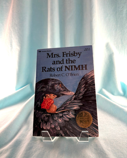 Book titled 'Mrs. Frisby and the Rats of NIMH' by Robert C. O'Brien on a stand against a light curtain background.