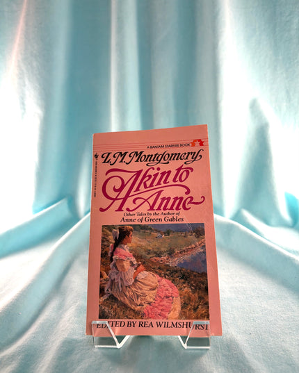Book titled 'Akin to Anne' by L.M. Montgomery on a stand against a light blue curtain background
