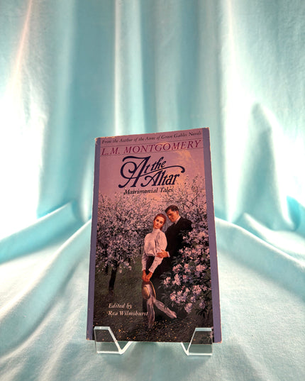 Book titled 'At the Altar' by L.M. Montgomery on a stand against a light blue curtain background