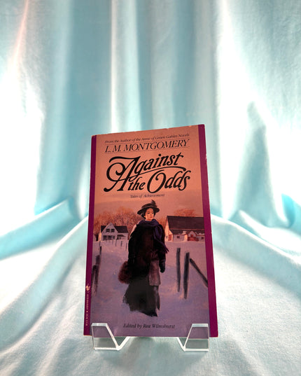 Book titled 'Against the Odds' by L.M. Montgomery on a stand against a light fabric background