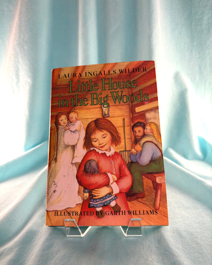 Book titled 'Little House in the Big Woods' by Laura Ingalls Wilder on a stand against a light background