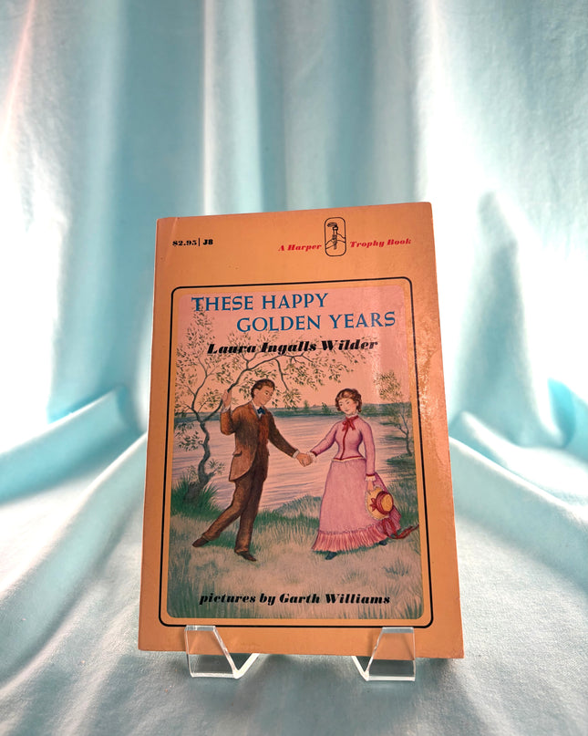 Book titled 'These Happy Golden Years' by Laura Ingalls Wilder on a stand with a light blue curtain background.