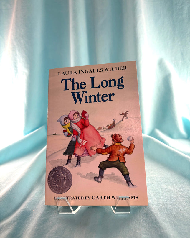 Book titled 'The Long Winter' by Laura Ingalls Wilder on a stand with a light blue curtain background