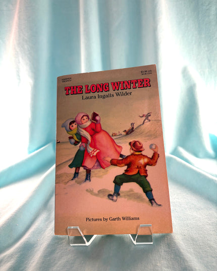 Book titled 'The Long Winter' by Laura Ingalls Wilder with illustrations on a light blue curtain background.