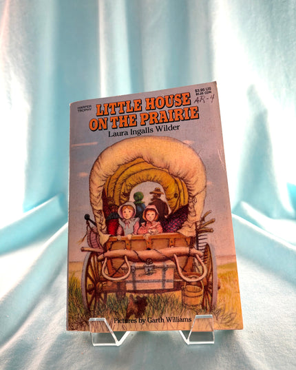 Book titled 'Little House on the Prairie' by Laura Ingalls Wilder on a stand against a light blue curtain background.