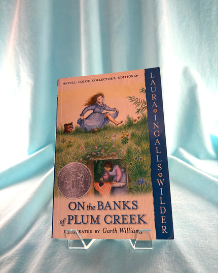 Book cover of 'On the Banks of Plum Creek' by Laura Ingalls Wilder on a light blue fabric background