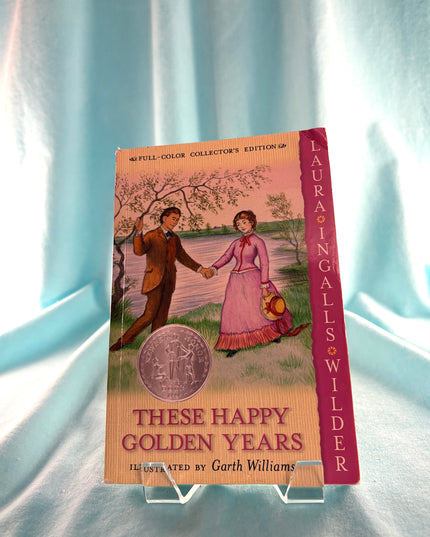 Book titled 'These Happy Golden Years' by Laura Ingalls Wilder on a stand with a light blue curtain background.