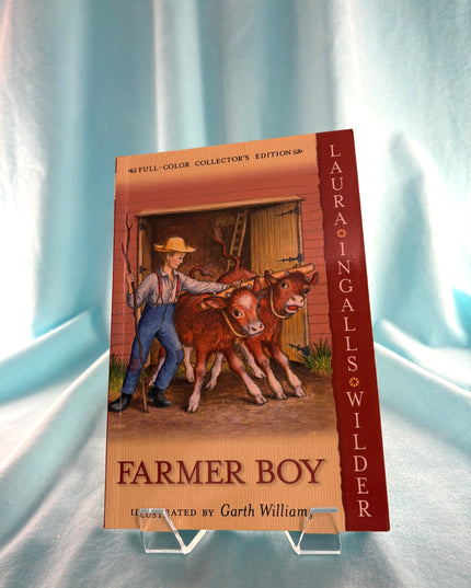 Book cover of 'Farmer Boy' by Laura Ingalls Wilder on a stand against a light blue curtain background
