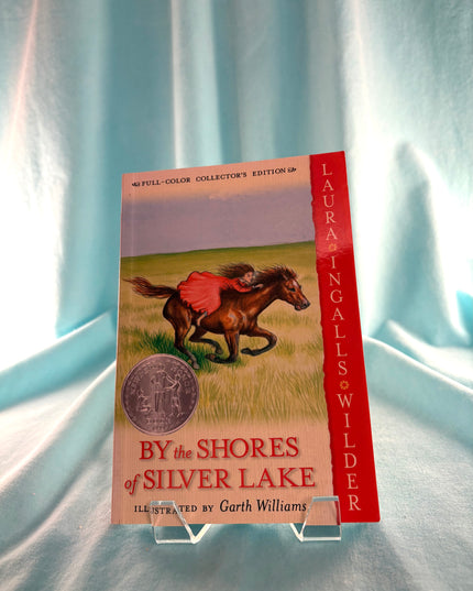 Book titled 'By the Shores of Silver Lake' by Laura Ingalls Wilder on a light blue fabric background