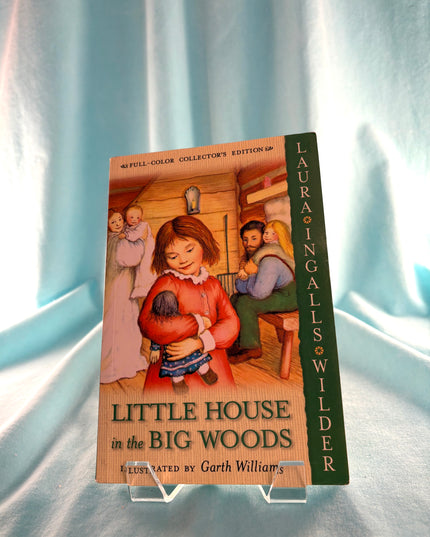 Book titled 'Little House in the Big Woods' by Laura Ingalls Wilder on a stand against a light blue curtain background.