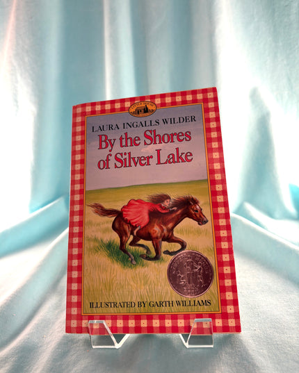 Book titled 'By the Shores of Silver Lake' by Laura Ingalls Wilder on a stand with a checkered border.