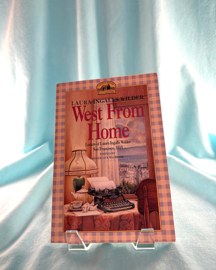 Book titled 'West From Home' by Laura Ingalls Wilder on a checkered stand against a light blue curtain background.